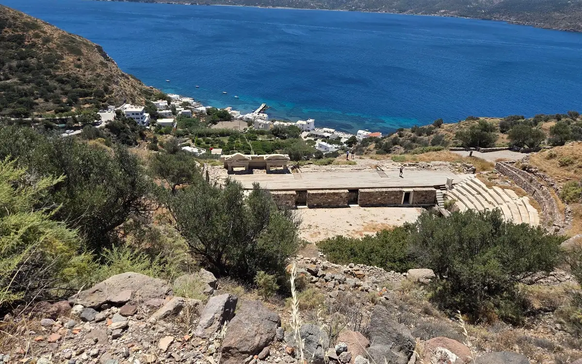 Steinstufen eines antiken Theaters zwischen Olivengebüsch, mit einem Dorf und einer tiefblauen Bucht darunter