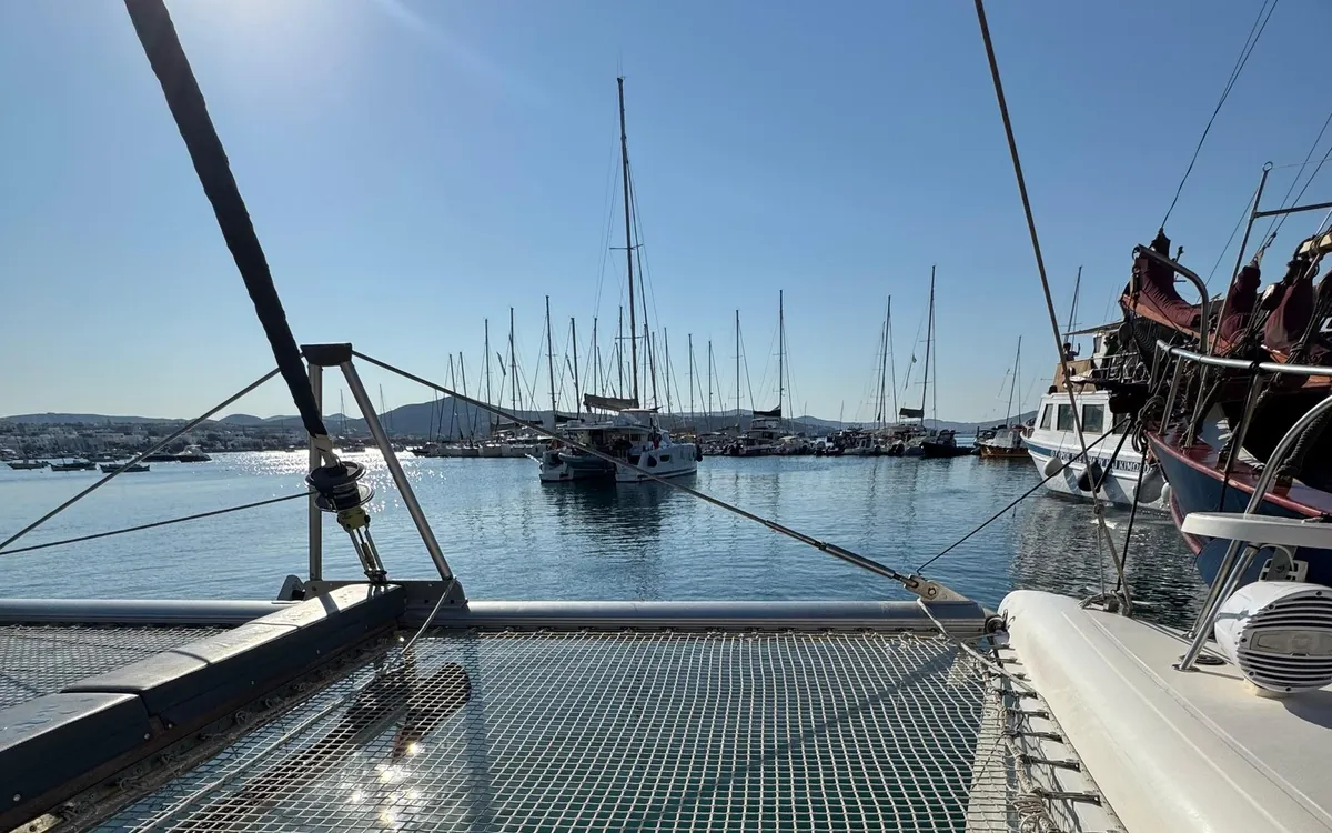 La red de un catamarán en primer plano, con una fila de mástiles de veleros en un puerto al frente