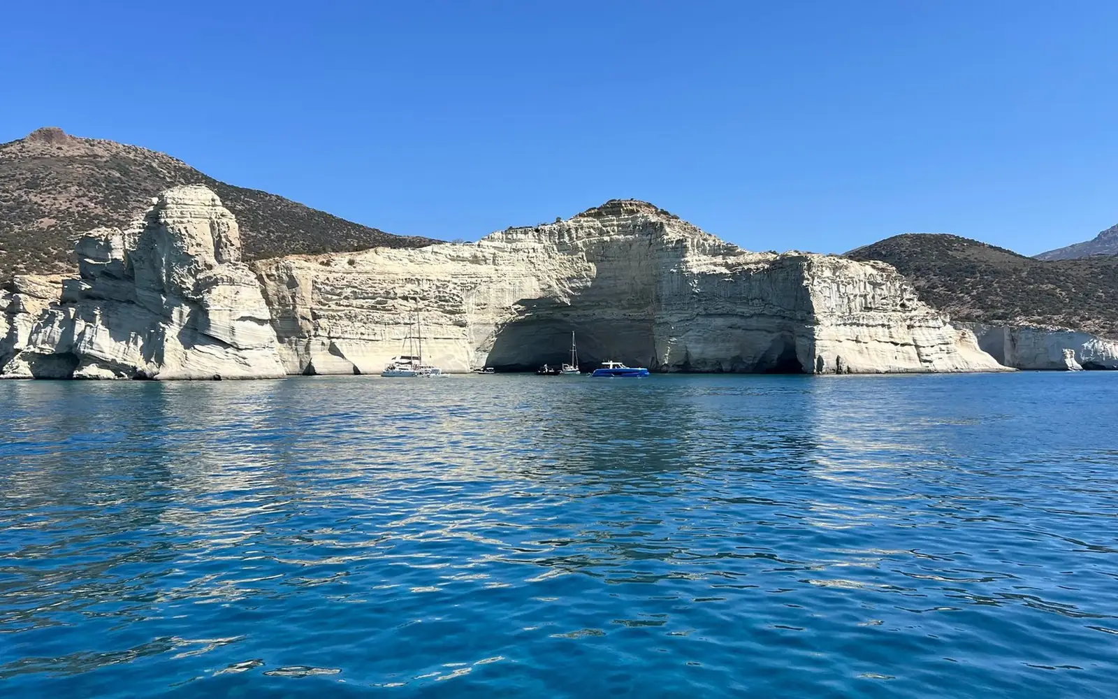 Weiße geschichtete Klippen mit flachen Meereshöhlen an der Küste von Milos, darunter ankernde Boote