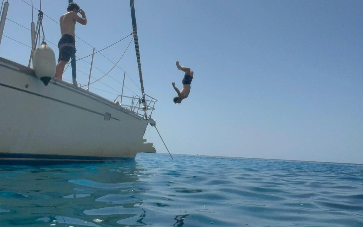 A swimmer flipping off the side of a sailboat into open blue water