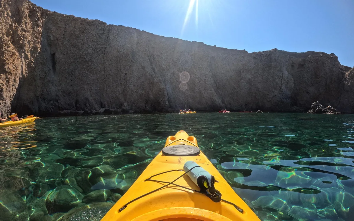 La proue jaune d'un kayak au-dessus d'une eau verte limpide, avec des kayakistes sous une falaise sombre
