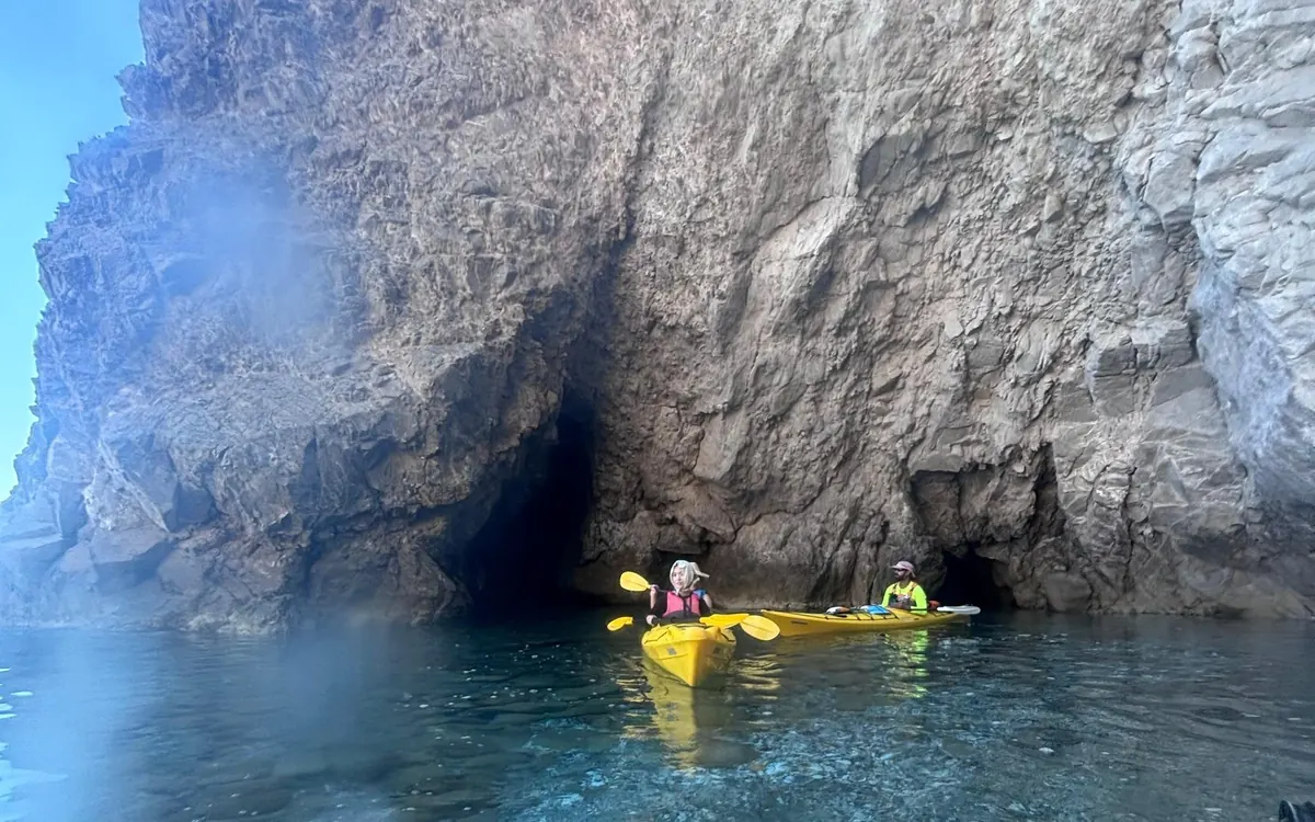 Zwei Personen in gelben Kajaks an der dunklen Öffnung einer Meereshöhle unter einer grauen Klippe