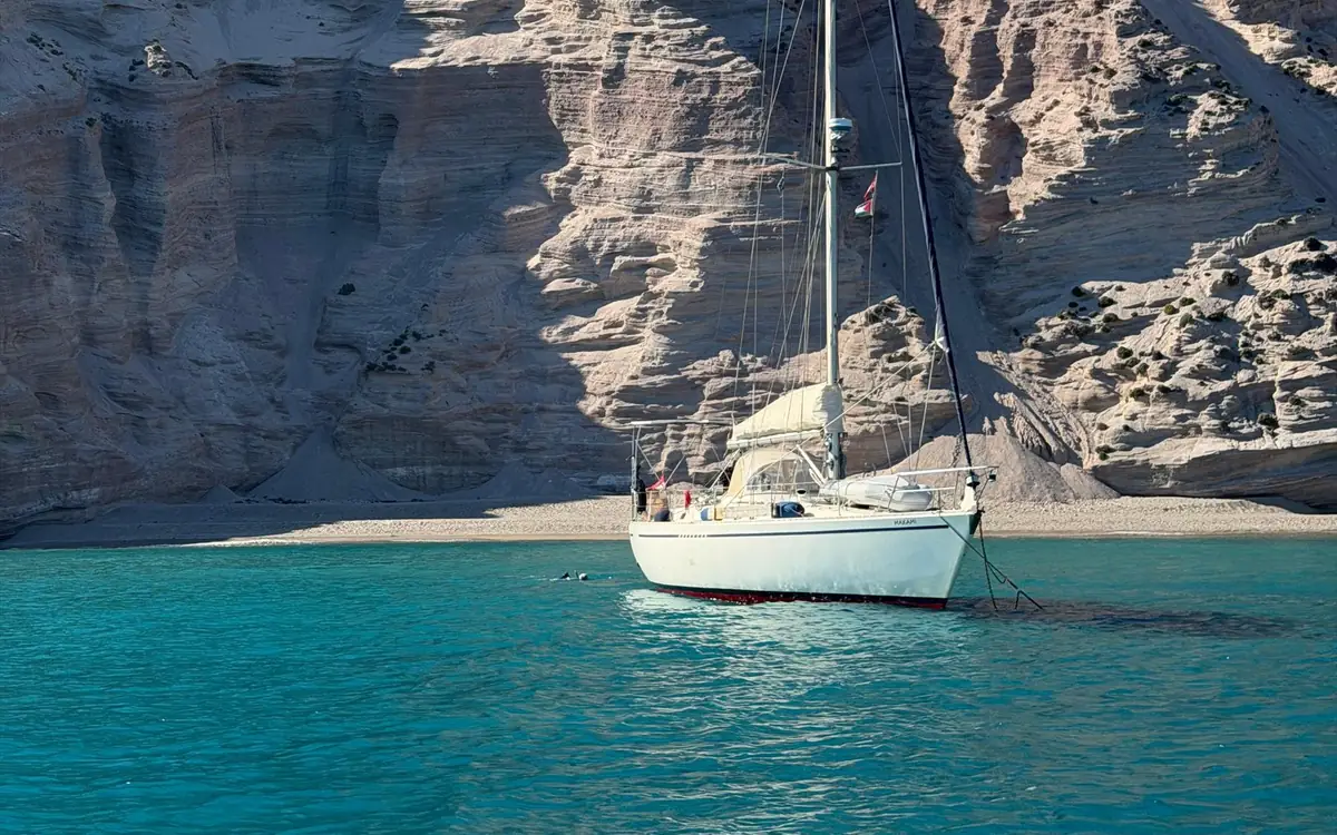 Un voilier blanc dans une eau turquoise au pied de hautes falaises pâles et stratifiées, avec une bande de plage grise