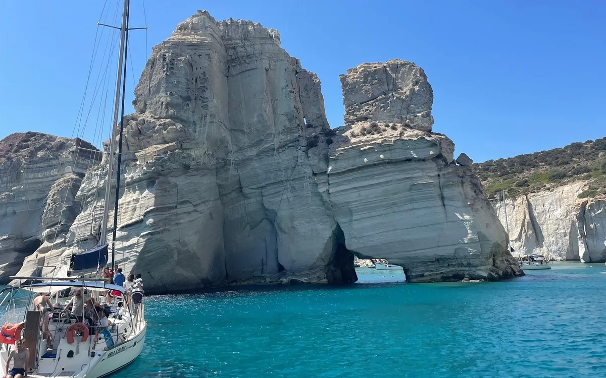 A sailboat with people on deck beside the white sea stacks and rock arch of Kleftiko