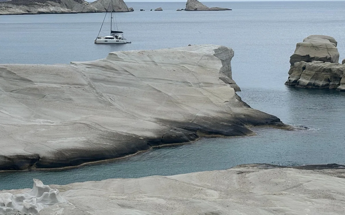 Glattes weißes Vulkangestein, das in grau-blaues Wasser übergeht, mit einem Segelboot vor der Küste